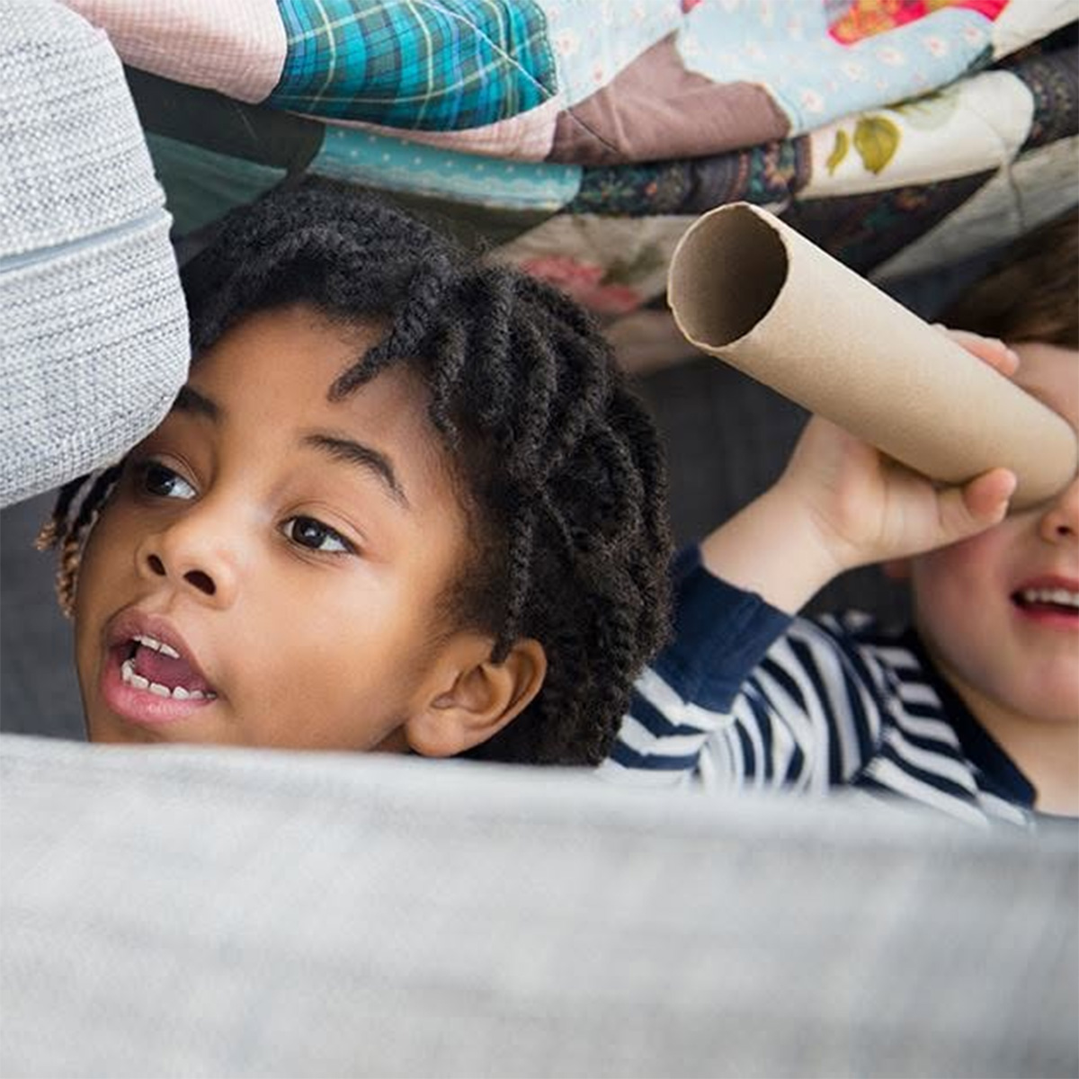 Two children looking through a cardboard tube while building a blanket fort
